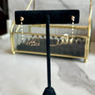 A pair of gold dangle earrings displayed on a black velvet jewelry stand, with a gold-framed jewelry organizer in the blurred background on a marble surface.
