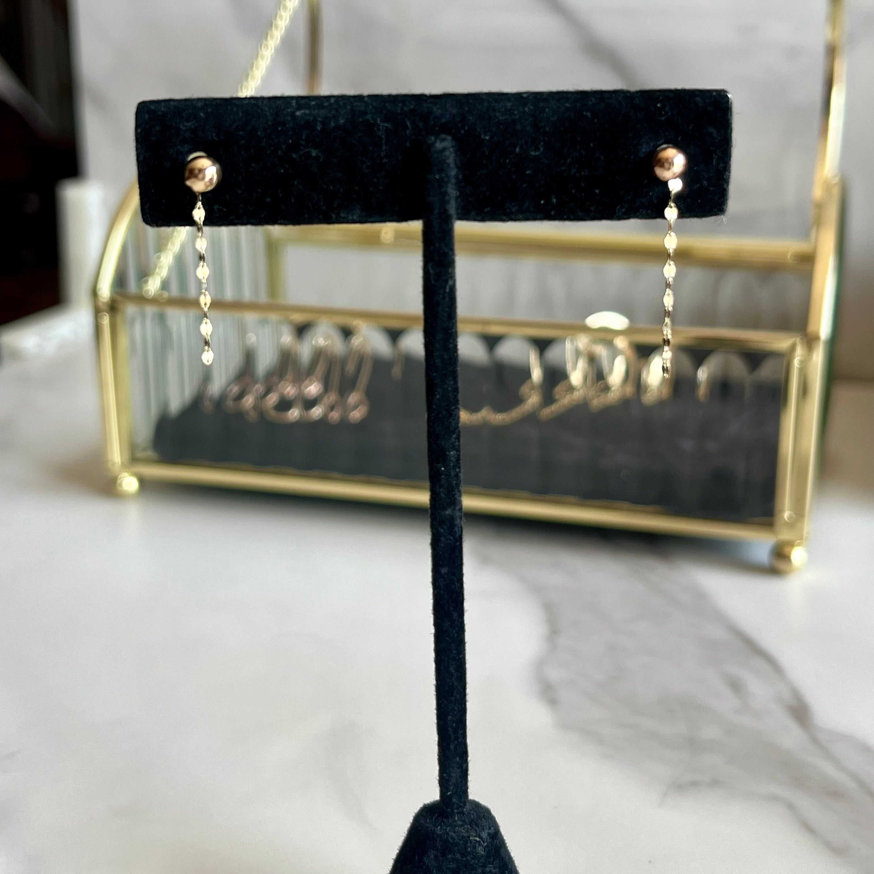 A pair of gold dangle earrings displayed on a black velvet jewelry stand, with a gold-framed jewelry organizer in the blurred background on a marble surface.