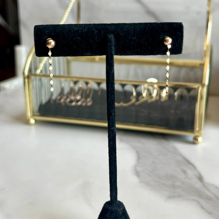 A pair of gold dangle earrings displayed on a black velvet jewelry stand, with a gold-framed jewelry organizer in the blurred background on a marble surface.