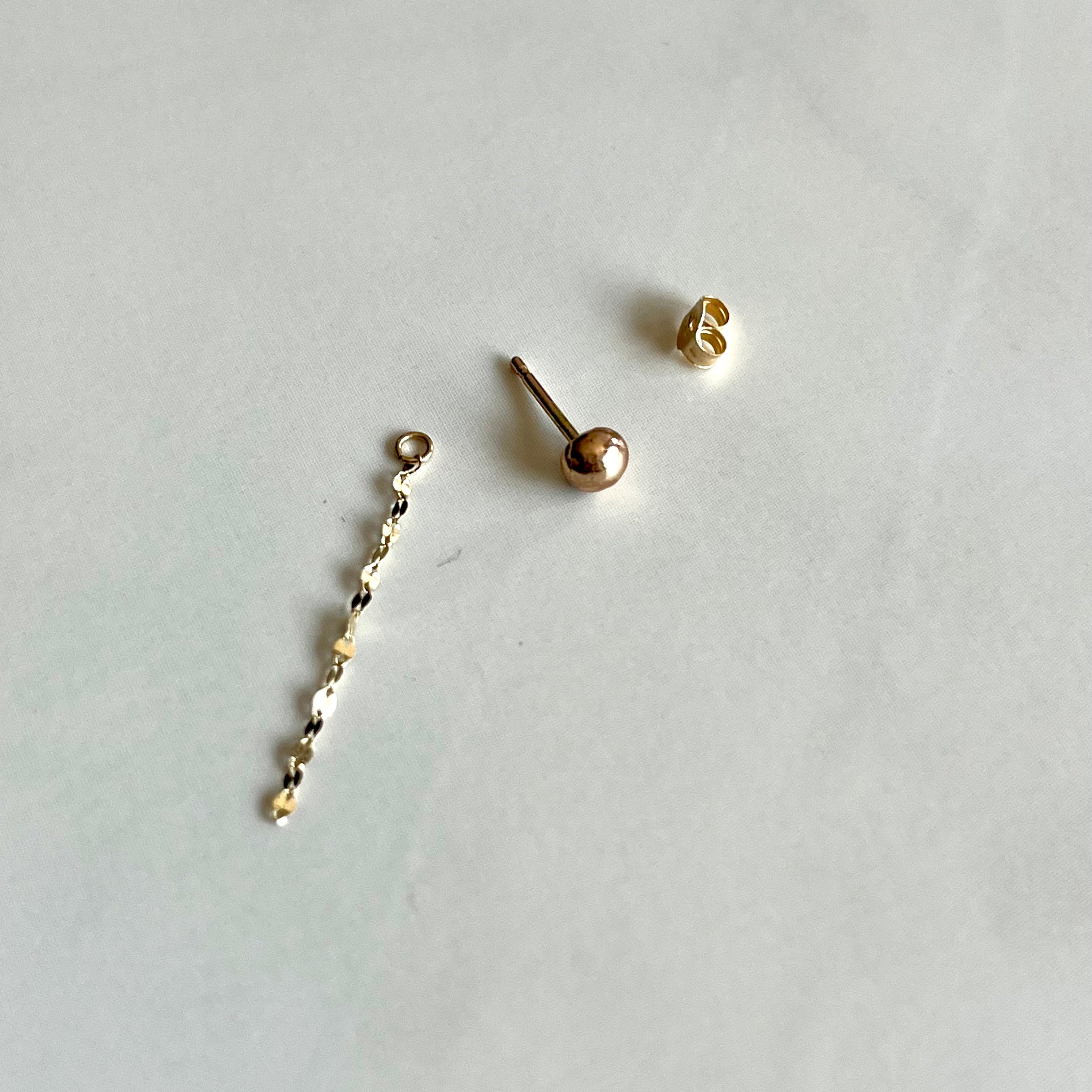 A pair of 14kt solid gold stud earrings shown disassembled on a light gray surface, with one earring showing the ball-shaped stud and the other showing the butterfly backing.